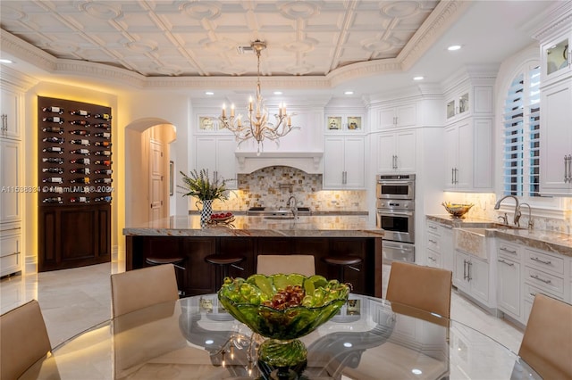 kitchen featuring a kitchen bar, light stone countertops, stainless steel double oven, decorative light fixtures, and a center island with sink