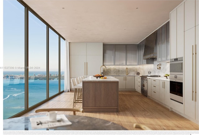 kitchen with an island with sink, floor to ceiling windows, wall chimney exhaust hood, a water view, and backsplash