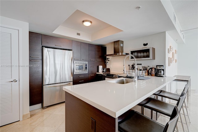 kitchen with a tray ceiling, appliances with stainless steel finishes, a kitchen bar, wall chimney range hood, and dark brown cabinets