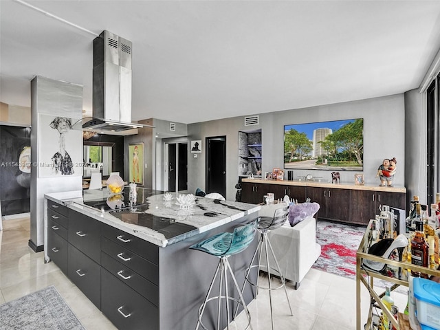 kitchen featuring wall chimney range hood, light tile flooring, light stone countertops, a breakfast bar, and black electric stovetop