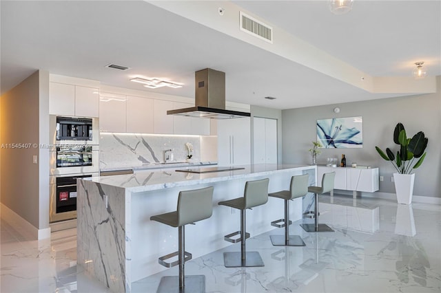 kitchen featuring island range hood, a kitchen breakfast bar, white cabinets, backsplash, and light stone counters