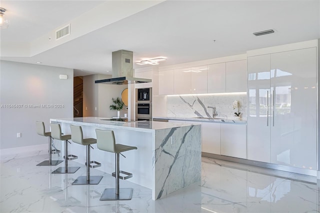 kitchen featuring a kitchen island with sink, oven, white cabinets, a breakfast bar, and island range hood