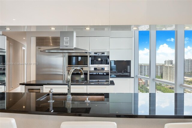 kitchen with dark stone counters, white cabinets, wall chimney exhaust hood, and double oven