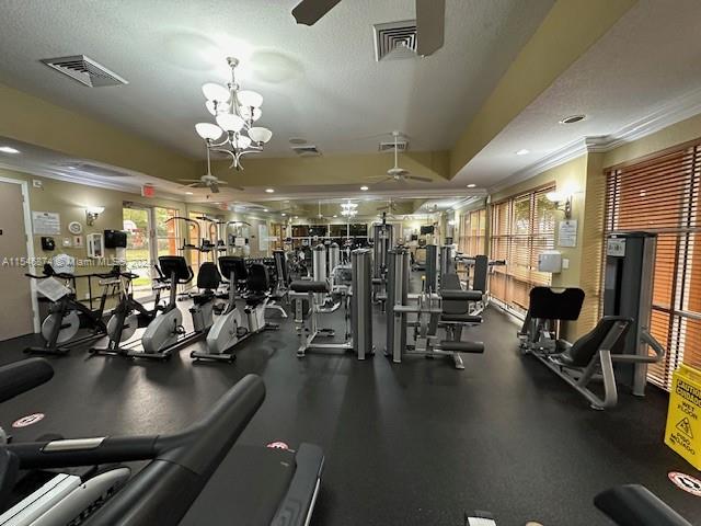 exercise room with crown molding, a textured ceiling, ceiling fan with notable chandelier, and a raised ceiling