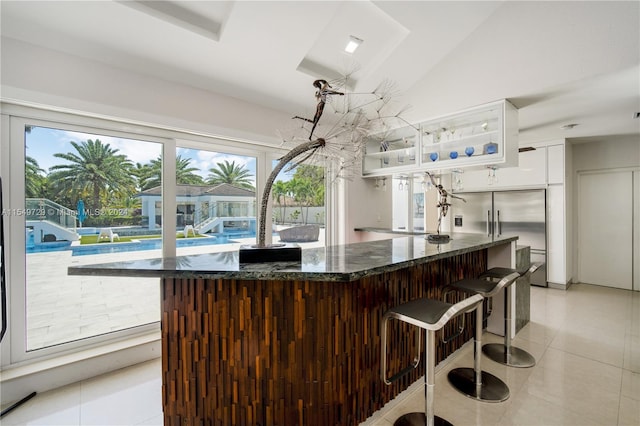 bar with stainless steel built in refrigerator, dark stone counters, white cabinetry, and light tile floors