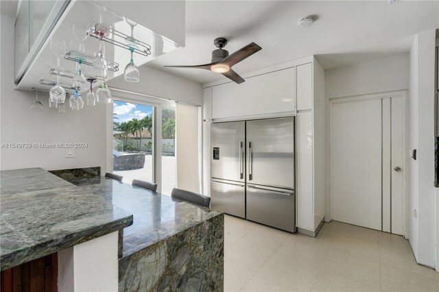 kitchen featuring built in refrigerator, ceiling fan, dark stone countertops, white cabinets, and light tile floors