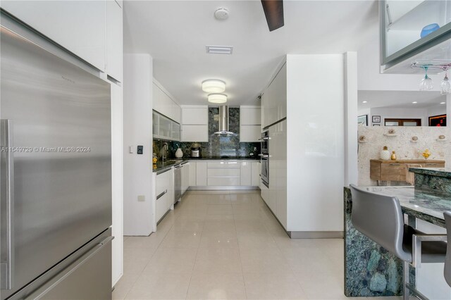 kitchen featuring light tile floors, white cabinets, tasteful backsplash, wall chimney range hood, and stainless steel appliances