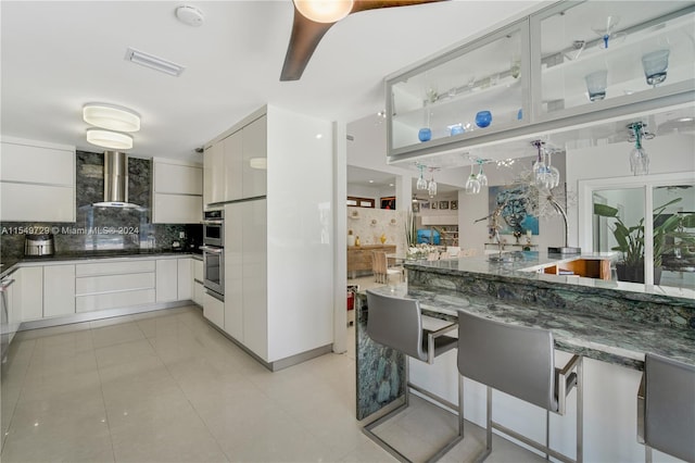 kitchen with light tile flooring, backsplash, wall chimney range hood, stainless steel double oven, and dark stone countertops