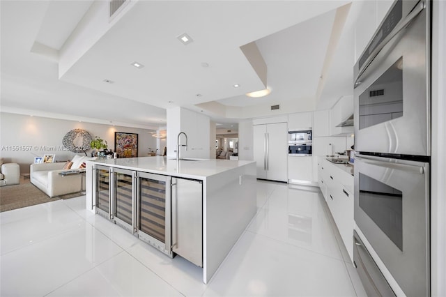 kitchen with built in appliances, light tile floors, white cabinets, a raised ceiling, and a kitchen island with sink