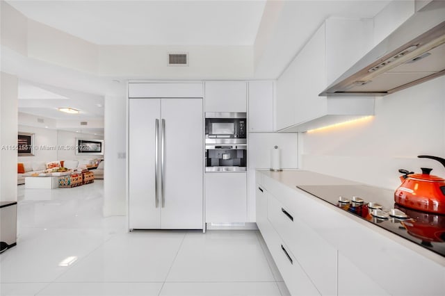 kitchen with built in appliances, a raised ceiling, white cabinets, light tile flooring, and wall chimney range hood