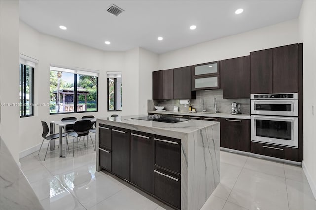 kitchen featuring light tile floors, black electric stovetop, stainless steel double oven, dark brown cabinetry, and light stone counters