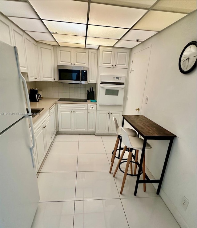 kitchen with light tile floors, a drop ceiling, tasteful backsplash, white appliances, and white cabinetry