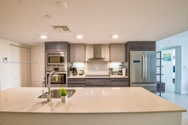 kitchen featuring sink, stainless steel appliances, light tile floors, a center island with sink, and wall chimney range hood