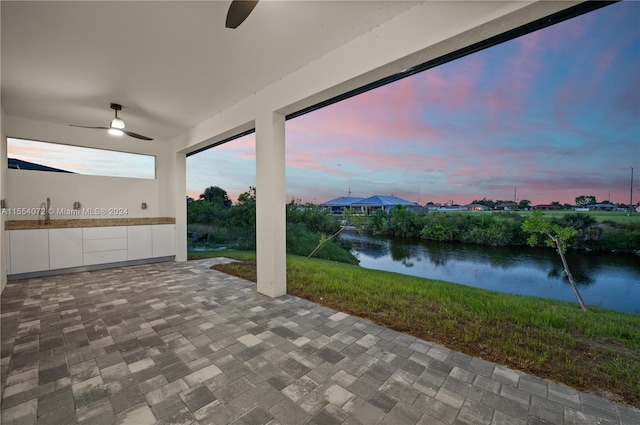 patio terrace at dusk with a ceiling fan, a water view, and a lawn