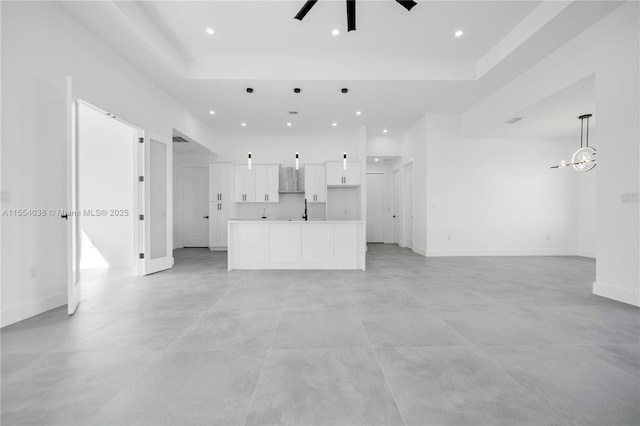 unfurnished living room with ceiling fan with notable chandelier, a tray ceiling, recessed lighting, and baseboards