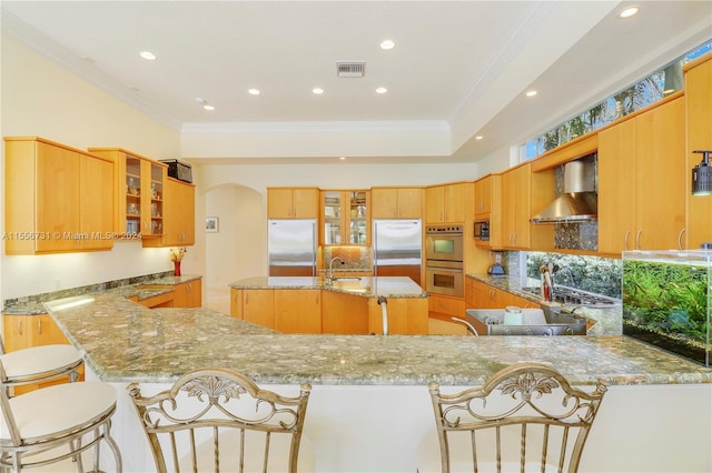 kitchen with appliances with stainless steel finishes, a breakfast bar, an island with sink, light stone counters, and wall chimney range hood