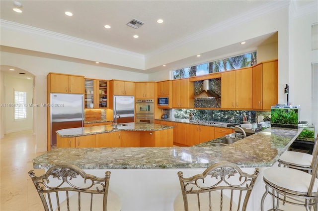 kitchen featuring a kitchen bar, sink, dark stone countertops, wall chimney exhaust hood, and built in appliances