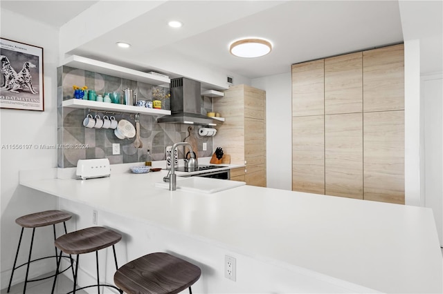 kitchen featuring backsplash, a kitchen bar, light brown cabinetry, and wall chimney range hood