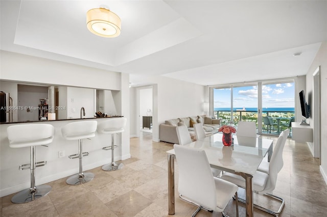 dining area featuring a water view, a raised ceiling, light tile floors, and a wall of windows