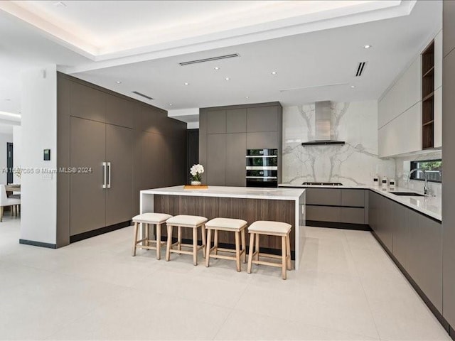 kitchen featuring wall chimney exhaust hood, tasteful backsplash, sink, light tile patterned floors, and a kitchen island