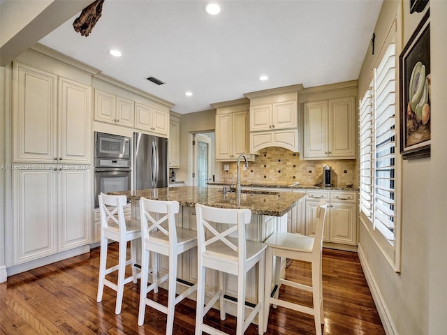 kitchen with stone counters, backsplash, dark wood-type flooring, a kitchen island with sink, and appliances with stainless steel finishes