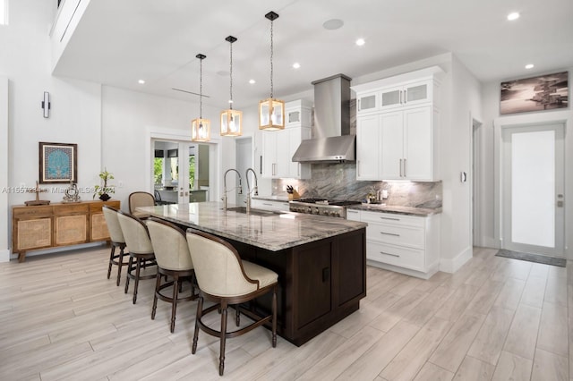 kitchen featuring pendant lighting, wall chimney range hood, light stone countertops, a kitchen island with sink, and sink