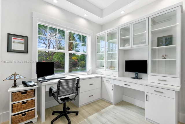 office space featuring a tray ceiling, built in desk, and light hardwood / wood-style flooring