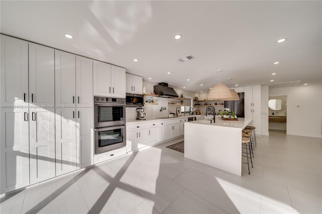 kitchen featuring stainless steel appliances, light tile floors, wall chimney exhaust hood, a kitchen island with sink, and white cabinetry