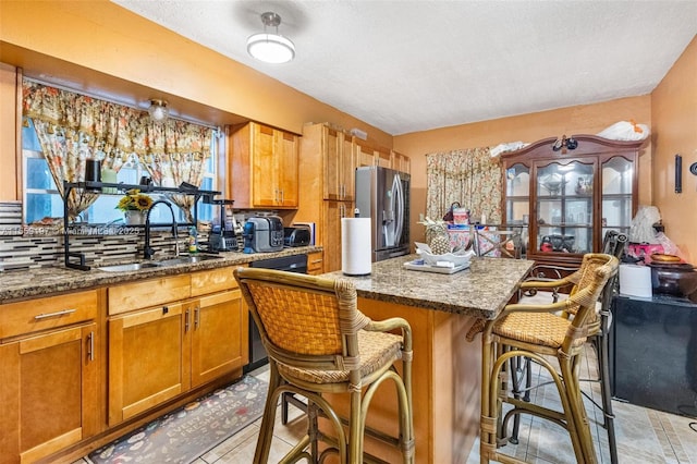 kitchen featuring stainless steel refrigerator with ice dispenser, brown cabinetry, a sink, dark stone countertops, and a kitchen breakfast bar