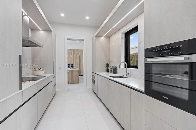 kitchen featuring stainless steel oven, wall chimney range hood, sink, black stovetop, and light tile patterned floors