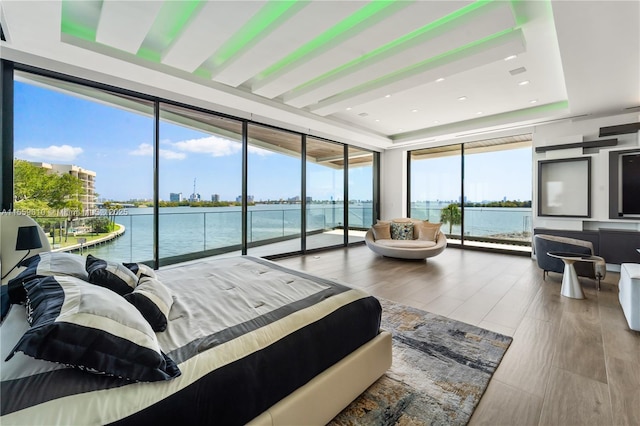 bedroom with wood-type flooring, a water view, a tray ceiling, a wall of windows, and access to outside