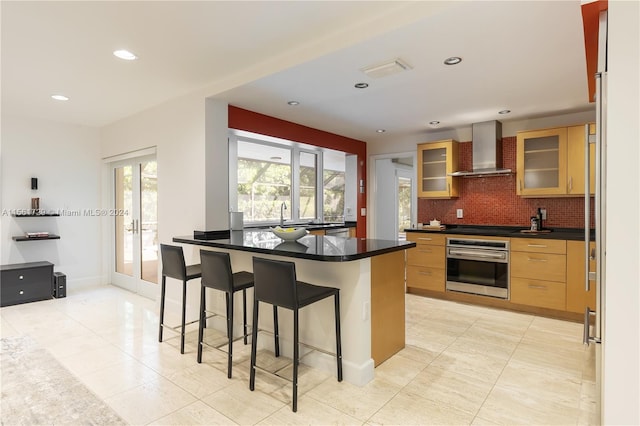 kitchen featuring wall chimney exhaust hood, a kitchen breakfast bar, stovetop, oven, and decorative backsplash