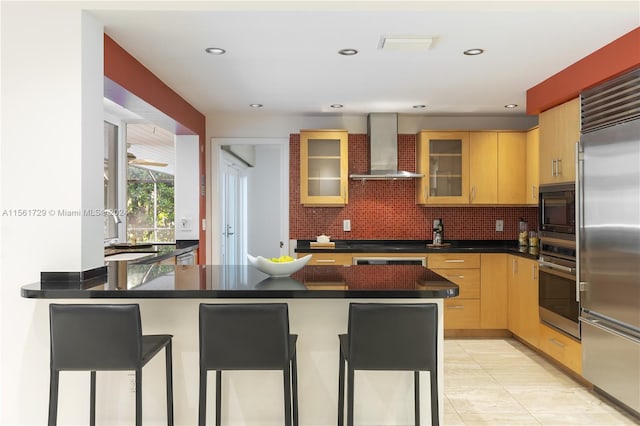 kitchen featuring a breakfast bar, built in appliances, backsplash, and wall chimney range hood