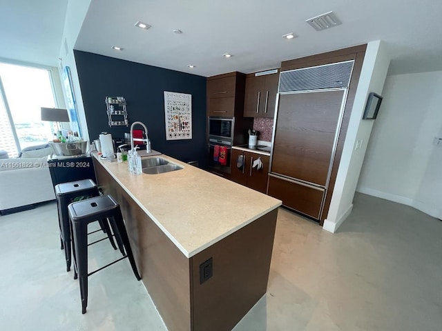 kitchen featuring a kitchen breakfast bar, built in refrigerator, dark brown cabinetry, a kitchen island with sink, and sink