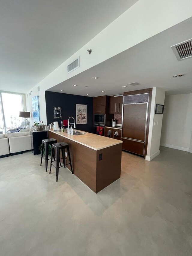 kitchen with built in appliances, a kitchen breakfast bar, dark brown cabinetry, a kitchen island with sink, and sink