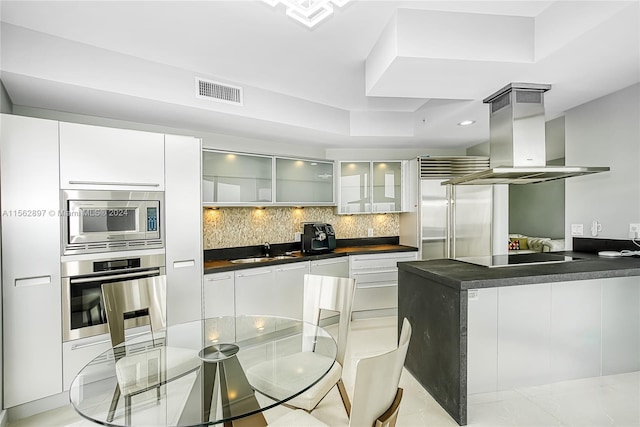 kitchen featuring built in appliances, light tile floors, backsplash, island range hood, and white cabinetry