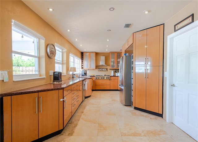 kitchen with wall chimney range hood, dark stone countertops, light tile flooring, and appliances with stainless steel finishes