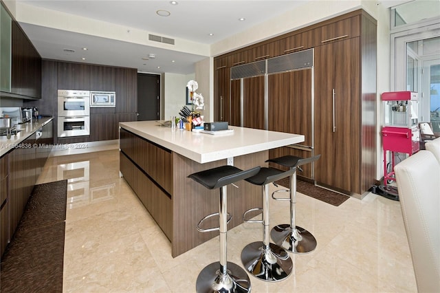 kitchen featuring a breakfast bar, a kitchen island, dark brown cabinets, built in appliances, and light tile flooring