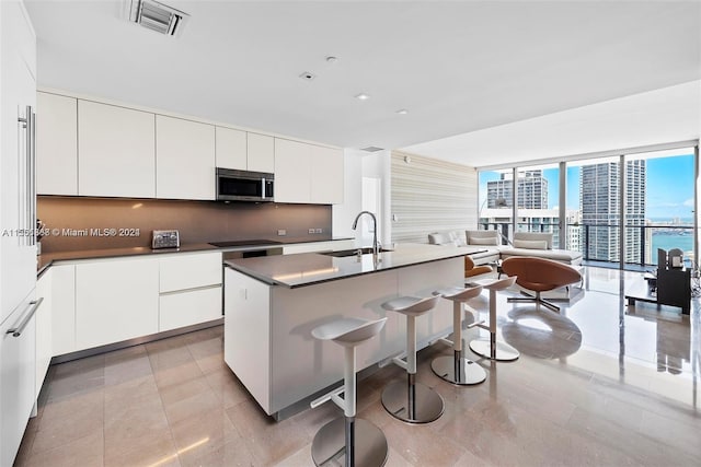 kitchen featuring an island with sink, a kitchen breakfast bar, floor to ceiling windows, white cabinets, and sink