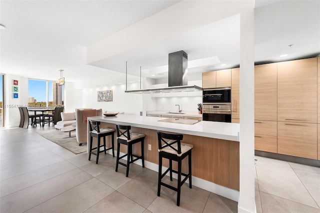 kitchen with light tile floors, light brown cabinetry, a breakfast bar area, island range hood, and stainless steel double oven