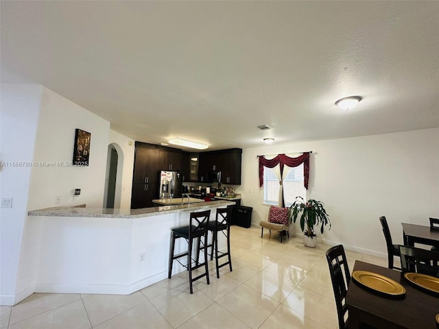 kitchen featuring a breakfast bar, dark brown cabinetry, light tile patterned flooring, stainless steel fridge with ice dispenser, and kitchen peninsula