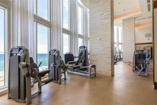 gym with a towering ceiling, a water view, a healthy amount of sunlight, and a tray ceiling