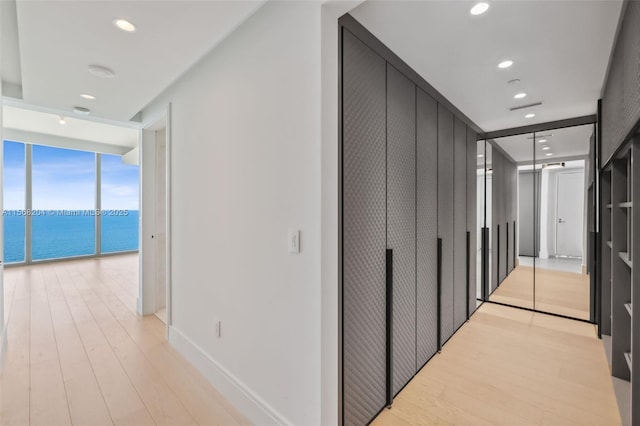 hall with light wood-type flooring, a water view, expansive windows, and recessed lighting