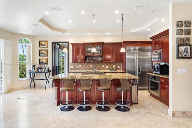 kitchen with pendant lighting, a breakfast bar, built in appliances, a tray ceiling, and light stone counters