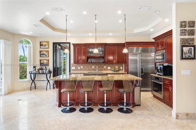 kitchen with decorative light fixtures, a kitchen bar, built in appliances, light stone counters, and a tray ceiling