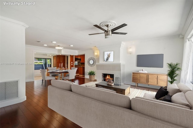 living room with ceiling fan, a tiled fireplace, dark hardwood / wood-style floors, and ornamental molding