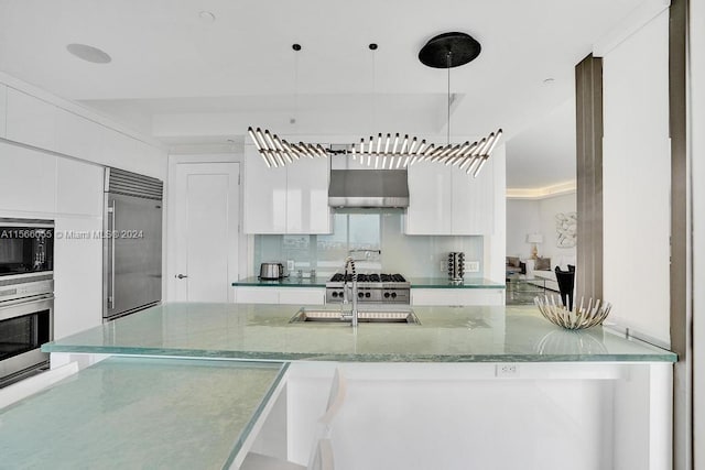 kitchen with built in appliances, wall chimney exhaust hood, white cabinets, and hanging light fixtures