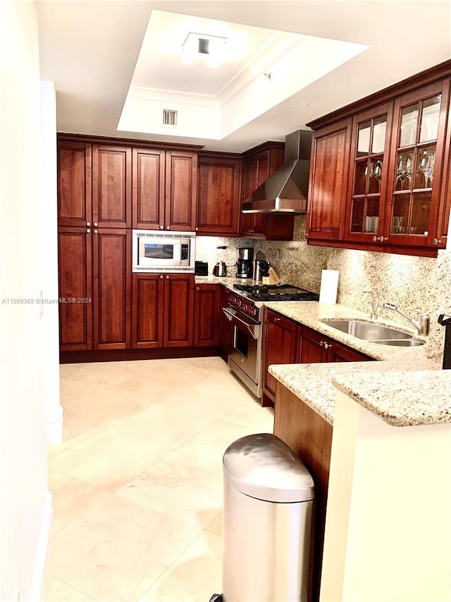 kitchen with stainless steel appliances, a tray ceiling, wall chimney range hood, light stone counters, and sink