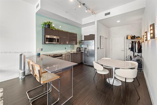 kitchen featuring dark wood-type flooring, dark brown cabinets, rail lighting, stainless steel appliances, and kitchen peninsula