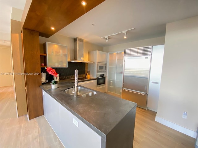 kitchen featuring built in appliances, kitchen peninsula, wall chimney exhaust hood, decorative backsplash, and sink